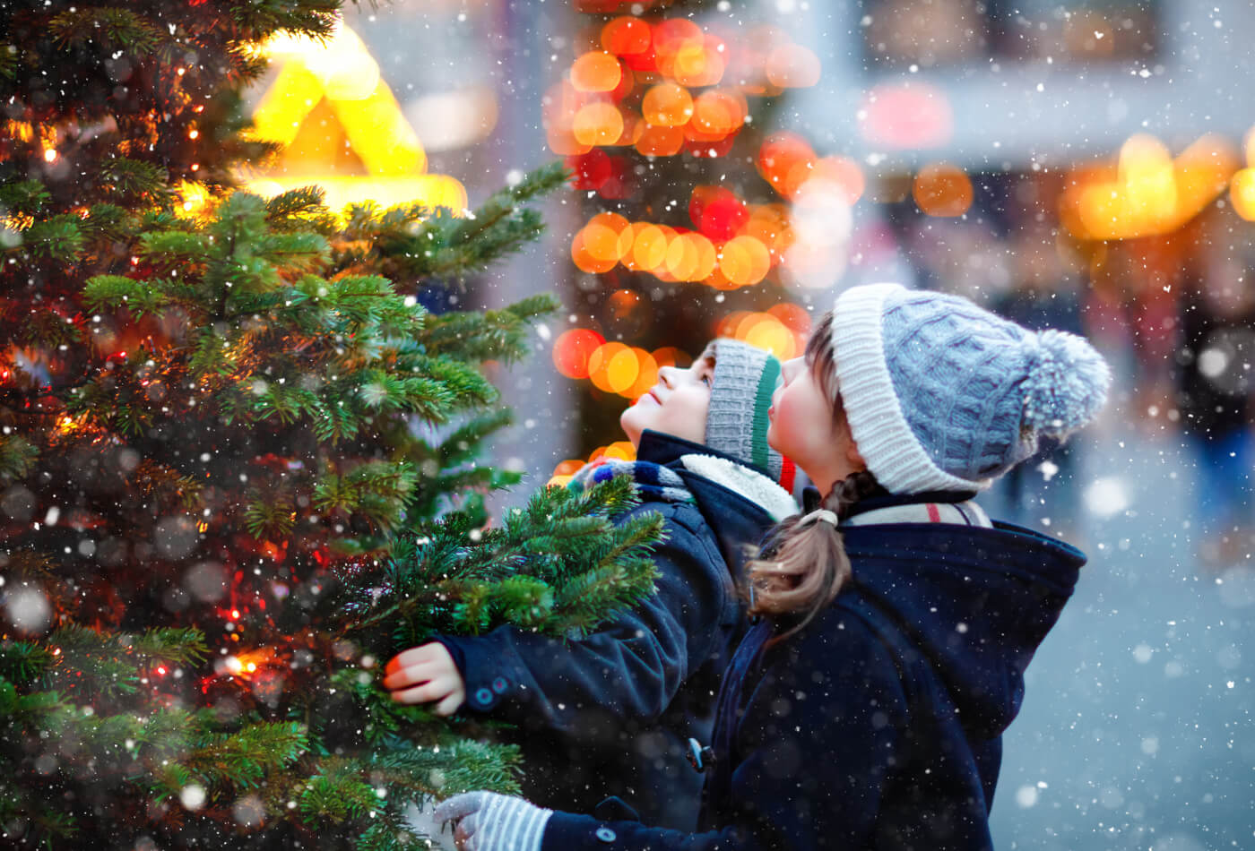 Two children looking up at a Christmas tree outside in the snow.