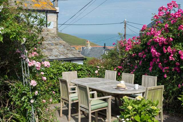 The garden of a North Cornwall holiday cottage, with a wooden table and chairs on patio and a sea view in the background.